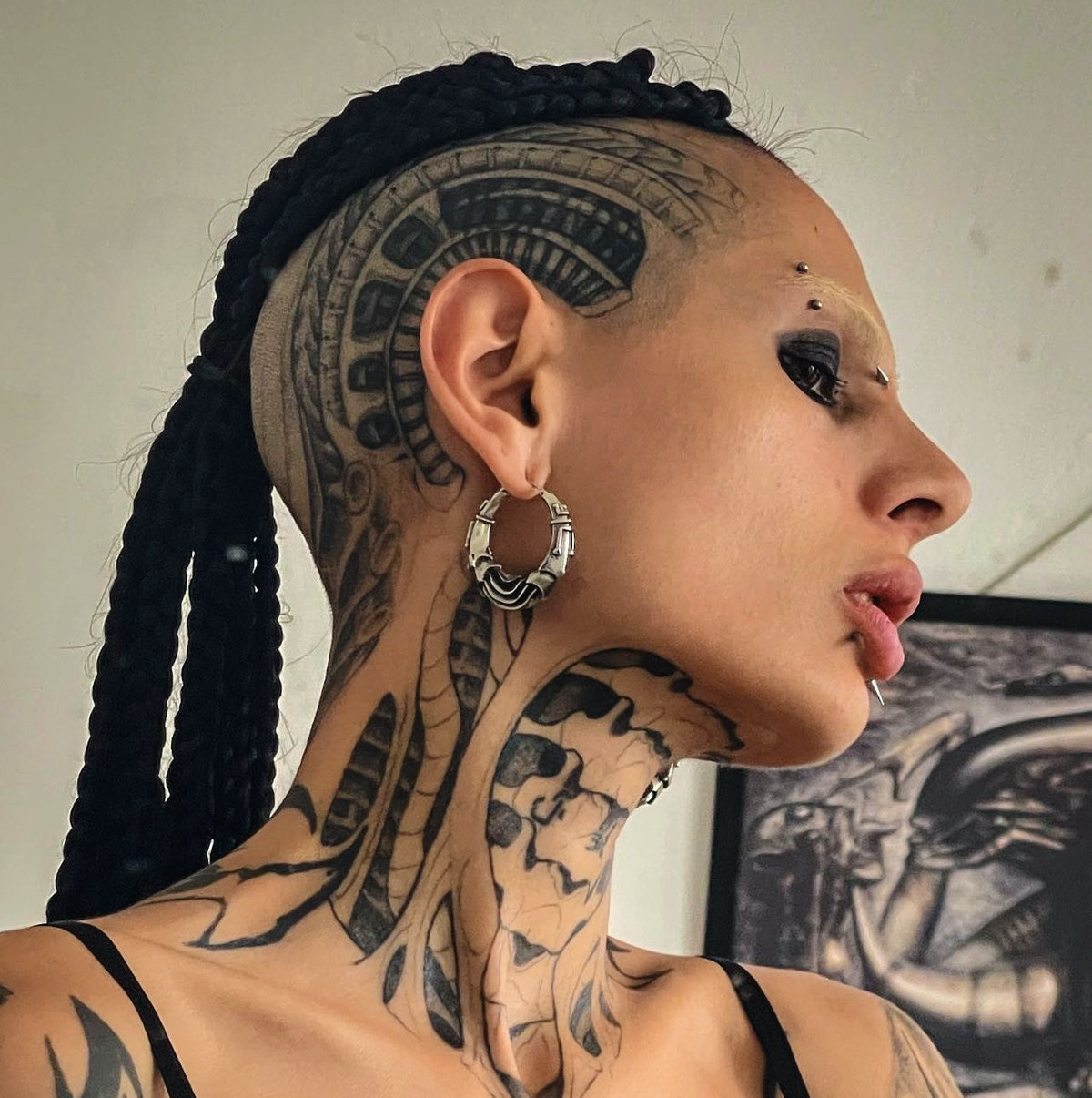 A person with intricate tattoos poses while wearing large circular metallic earrings, a septum ring, and black nail polish. Their dramatic eye makeup and red lighting create a bold and intense atmosphere. They touch their lips with a confident gaze.