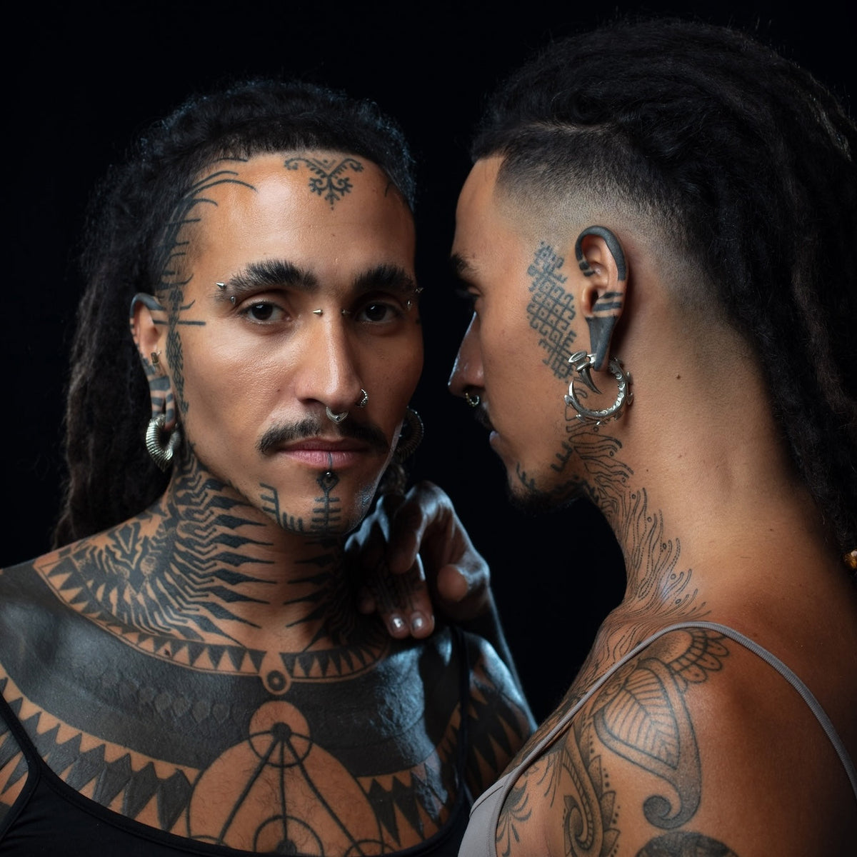 Two individuals with bold tribal facial and body tattoos face each other in a reflective pose. Both have septum piercings, eyebrow jewelry, and large hoop earrings, with one showing side and back details of their intricate tattoos and dreadlocks.