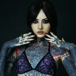 A heavily tattooed model with dark hair wearing a futuristic gold septum clicker. The intricate cyberpunk-inspired piercing is complemented by gothic rings, elongated nails, and bold makeup.