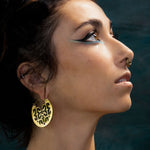 Artistic close-up of a woman looking upward, highlighting her turquoise eyeliner, bold black wing, and a detailed gold septum ring with subtle red gemstone accents. Her skin glows under soft lighting.