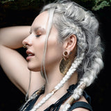 A woman with platinum blonde hair styled in intricate braids poses with her eyes closed and hand behind her head. She wears large, ornate earrings and an AYA Septum Clicker Shipibo Pattern by Kaya Movement. The background is dark with a hint of greenery at the top.