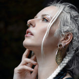 A woman with long, braided white hair stands in profile, gazing upwards with a thoughtful expression. She wears large, intricate earrings and an AYA Septum Clicker Shipibo Pattern Inspired by icaros patterns Gold from Kaya Movement. One hand gently touches her face. The background is dark and out of focus, highlighting her face and accessories.