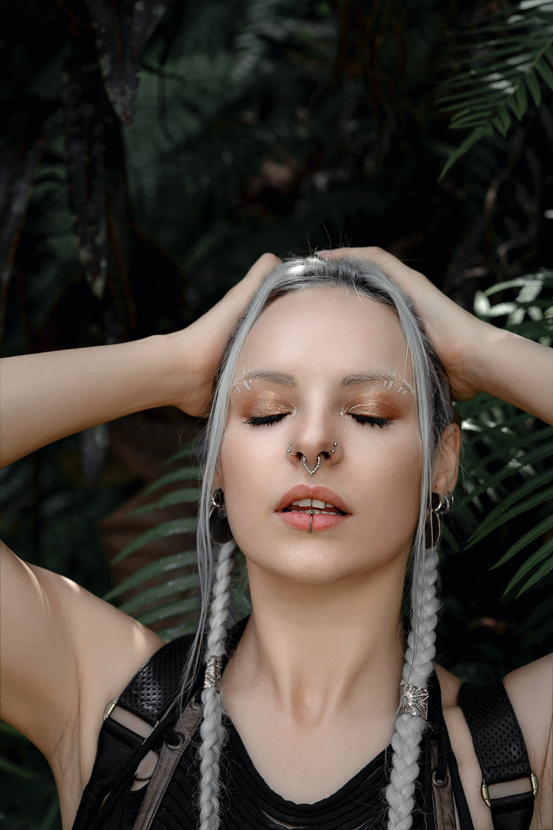 A person with closed eyes stands with their hands on their head. They have long, silver hair in braids, facial piercings including a KAMO Biomech Teardrop Clicker Septum Ring in Silver by Kaya Movement, and wear dark clothing. A lush, green background with foliage surrounds them.