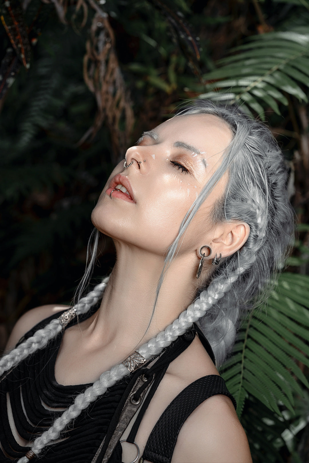 A person with long, grey braided hair and dark eye makeup is standing outdoors against a backdrop of green foliage. They are adorned with multiple earrings, including the Kaya Movement KAMO Biomech Teardrop Clicker Septum Ring in Silver (16 gauge) and stud earrings, while their eyes are closed and their head is tilted slightly back.