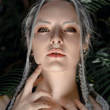 A person with light skin and blonde braids gazes intensely at the camera. They have facial piercings, including a striking Kaya Movement KAMO Biomech Teardrop Clicker Septum Ring in Silver, 16 gauge, and wear edgy makeup that highlights their cheekbones. Their hands lightly touch their chin and collarbone. Lush greenery forms the background.