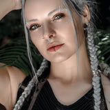 A woman with long, braided silver hair is looking at the camera, showcasing her KAMO Biomech Teardrop Clicker Septum Ring in sterling silver by Kaya Movement and several earrings. Wearing a black, textured top and handcrafted body jewelry, she poses with her hand resting on her head against a backdrop of green foliage.