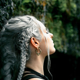 A woman with long, braided, silver hair and gold MARVDASHT Small Cyborg Hoop Clicker Ear Weights in 2 gauge by Kaya Movement stands in profile against a backdrop of lush greenery and rocks. She is looking upward with a calm expression, dressed in a black top, and the natural light highlights her hair and face.