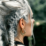 A person with long, silver braided hair is seen in profile. They are wearing large, ornate MARVDASHT Small Cyborg Hoop Clicker Ear Weights in Gold by Kaya Movement, and a dark sleeveless top. The background is blurred greenery, suggesting an outdoor setting.