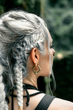 A person with long, silver braided hair is seen in profile. They are wearing large, ornate MARVDASHT Small Cyborg Hoop Clicker Ear Weights in Gold by Kaya Movement, and a dark sleeveless top. The background is blurred greenery, suggesting an outdoor setting.
