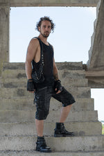 Full-body view of a model wearing a black leather double pouch holster bag, paired with black cargo shorts, combat boots, and a mesh tank top, standing on concrete steps.