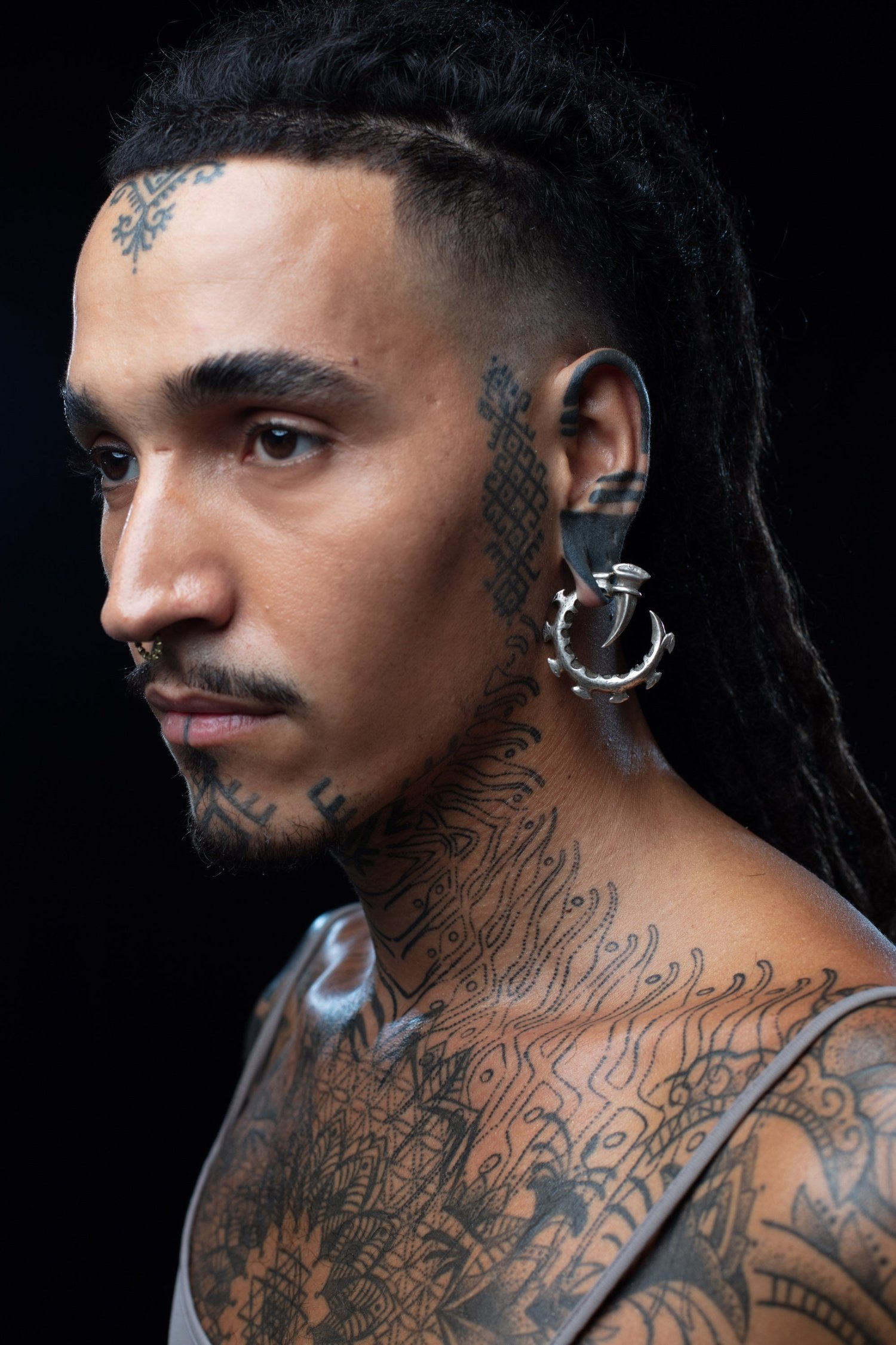 Profile portrait showing detailed facial tattoos and a silver biomechanical hoop ear weight. Subject has long dark dreadlocks, facial piercings, and extensive neck/shoulder tattoos featuring geometric patterns. Shot against dark background.