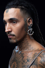 Profile portrait showing detailed facial tattoos and a silver biomechanical hoop ear weight. Subject has long dark dreadlocks, facial piercings, and extensive neck/shoulder tattoos featuring geometric patterns. Shot against dark background.