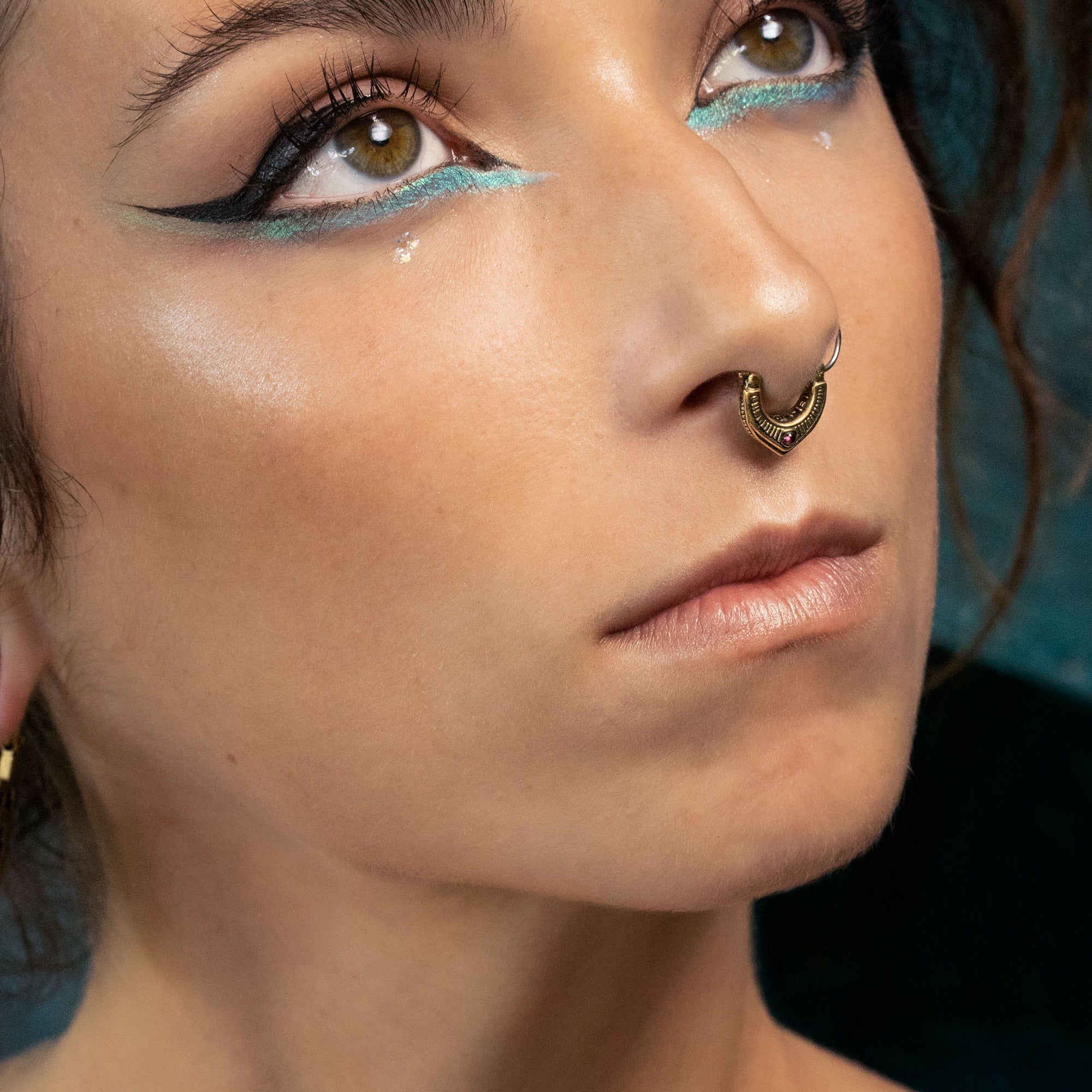 Artistic close-up of a woman looking upward, highlighting her turquoise eyeliner, bold black wing, and a detailed gold septum ring with subtle red gemstone accents. Her skin glows under soft lighting.
