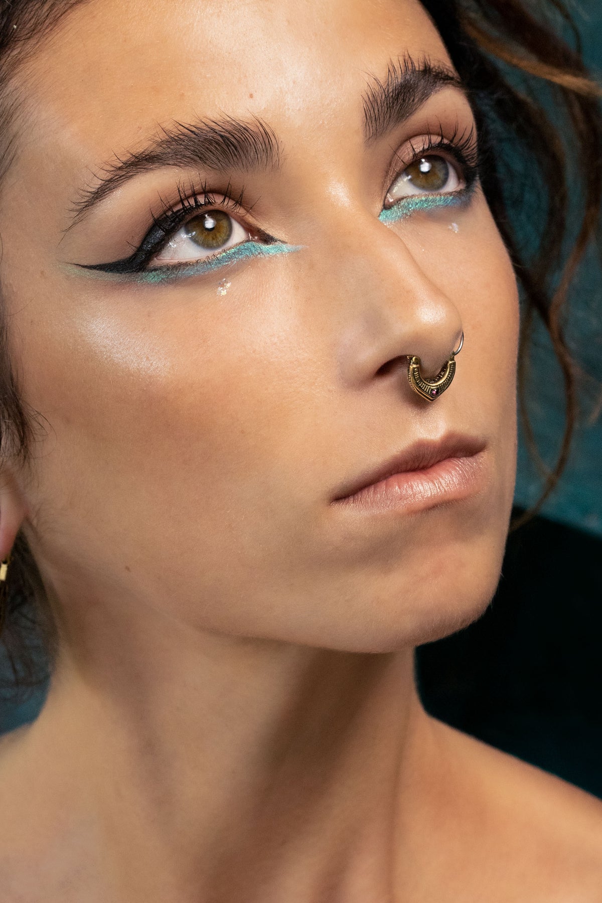 Artistic close-up of a woman looking upward, highlighting her turquoise eyeliner, bold black wing, and a detailed gold septum ring with subtle red gemstone accents. Her skin glows under soft lighting.