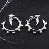 The GIGER Biomechanical Open Ended Hoop Ear Weights in Silver 925 from Kaya Movement are designed to resemble curved claws gripping an intricate, spiked ring. The jewelry rests on a dark, textured surface with hints of white threads at the edges, accentuating the contrast between the metal and the background.