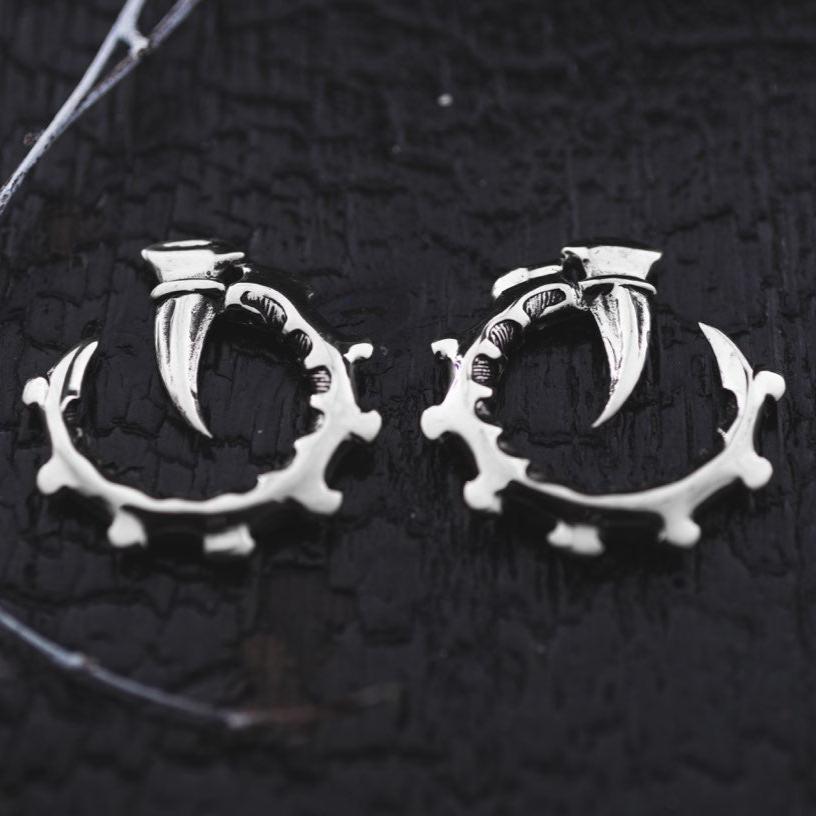 The GIGER Biomechanical Open Ended Hoop Ear Weights in Silver 925 from Kaya Movement are designed to resemble curved claws gripping an intricate, spiked ring. The jewelry rests on a dark, textured surface with hints of white threads at the edges, accentuating the contrast between the metal and the background.