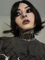 Close-up portrait of a tattooed woman with intense eye makeup, a cybernetic gold septum ring, and a bold black choker. Her alternative style embodies a dark futuristic aesthetic.