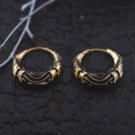 Close-up shot of a pair of Kaya Movement's PROMETHEUS Small Cyborg Hoop Earrings in Gold, 18 gauge, featuring an intricate textural design, set against a dark, charred wooden background. A small, delicate branch with slender twigs is visible to the left of the cyborg earrings, giving them a post-apocalyptic body jewelry vibe.