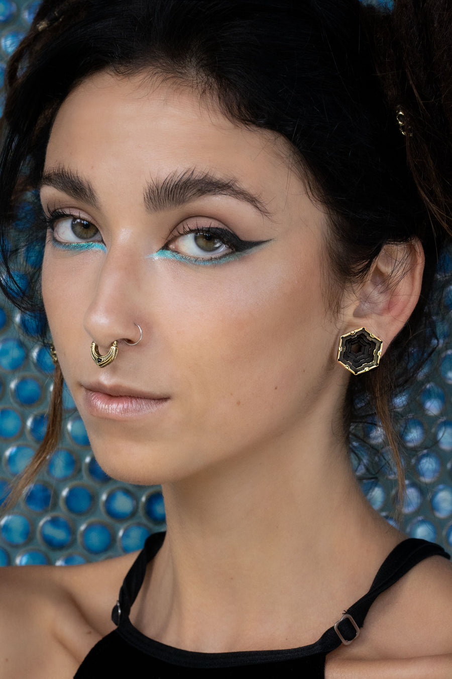 Portrait of a woman with turquoise and black winged eye makeup, wearing a detailed gold septum ring and matching hexagonal black-and-gold earrings. She gazes confidently at the camera.