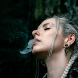 A person with light-colored braided hair and multiple ear piercings, including Kaya Movement's MARVDASHT Small Cyborg Hoop Clicker Ear Weights in Gold (2 gauge), blows smoke from their mouth. They have a nose ring and are wearing dark clothing. The background is dark and blurred, emphasizing the person.