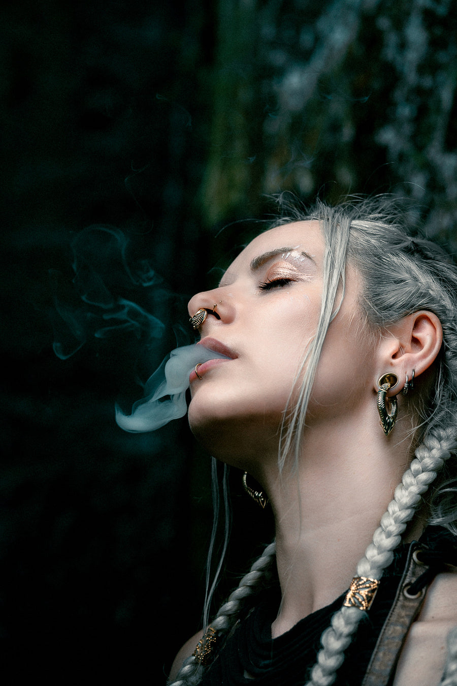 Woman exhaling smoke with closed eyes, wearing gold Xenomorph ear weights, emphasizing their fit and style.