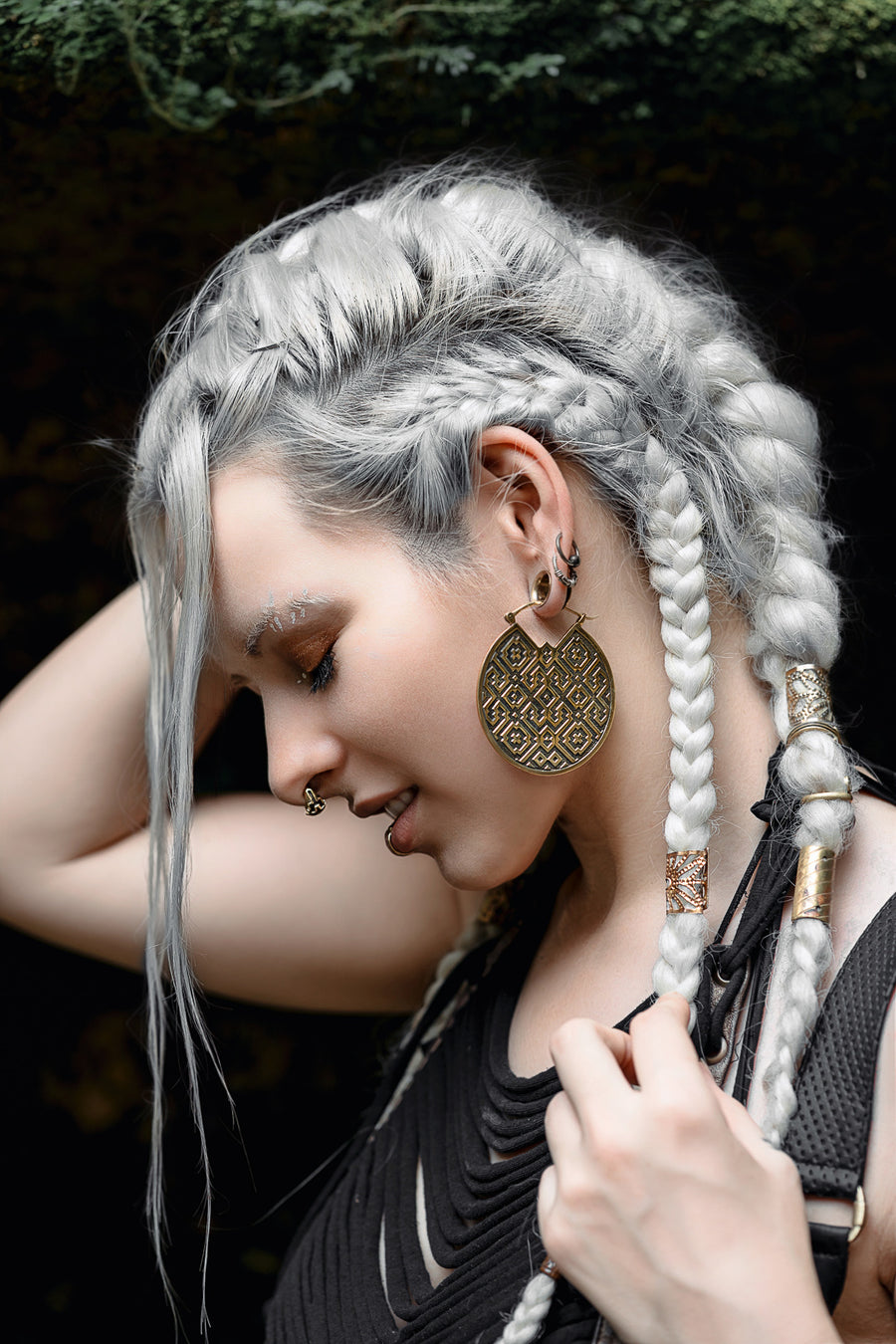 A profile view of a woman with silvery white hair styled in braids and loose strands, wearing ornate, disc-shaped earrings with a complex geometric pattern. She has a septum piercing and multiple ear piercings, subtly highlighted by her poised head tilt and gentle hand placement near her neck. The lighting casts a soft glow on her face, emphasizing her reflective expression