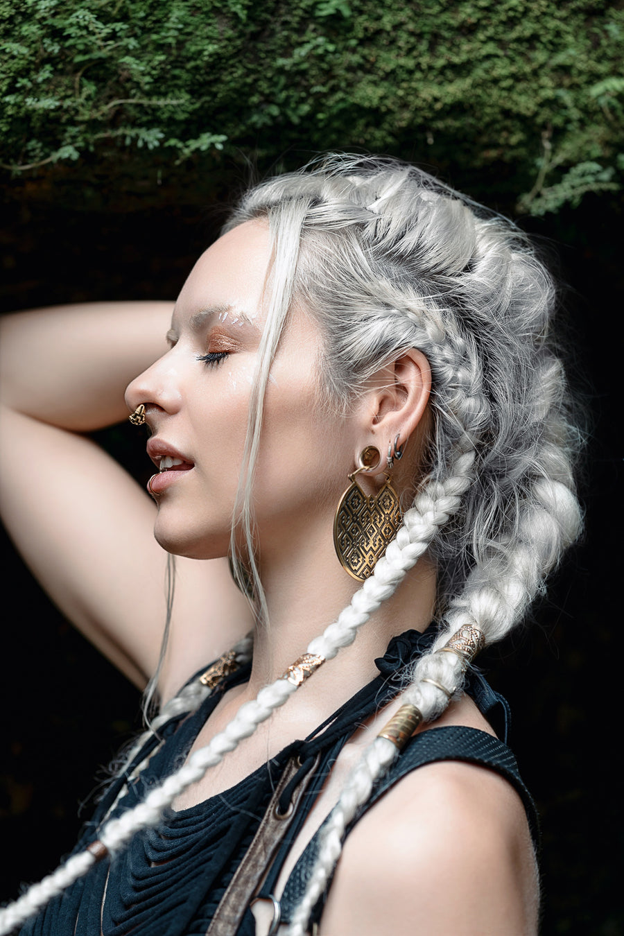 A serene profile of a woman with silver braided hair, exuding a bohemian vibe with her large, patterned, gold earrings. A sense of quiet confidence is conveyed through her closed eyes and the gentle placement of her hand on her head, as she stands against a natural backdrop that softly blurs into the distance.