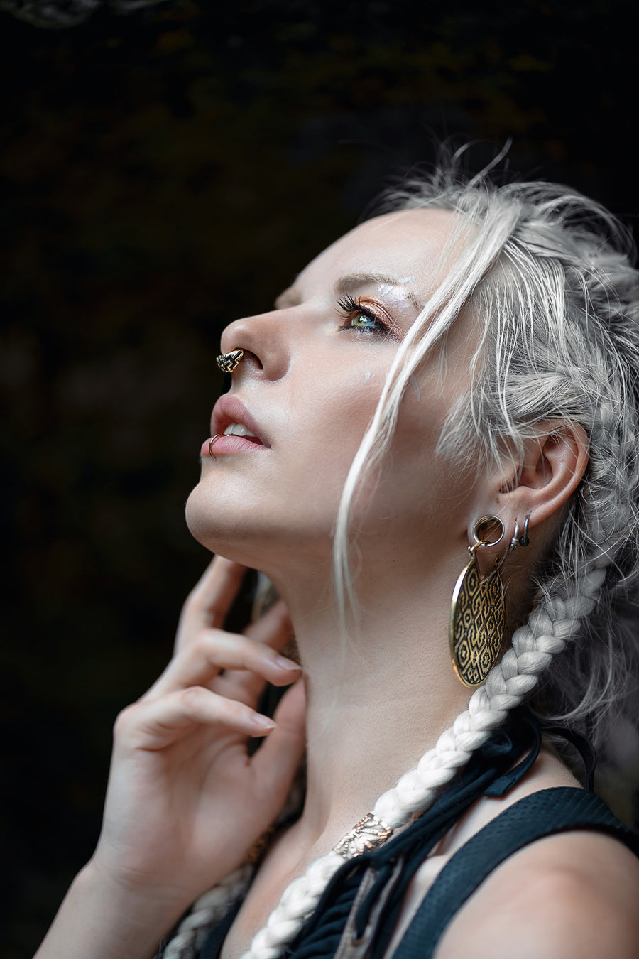 An image capturing a woman in a thoughtful pose, her gaze lifted skyward. She showcases intricate, disc-shaped earrings adorned with a detailed pattern, matched with a delicate septum ring. Her white hair, interwoven with braids and loose strands, cascades alongside her visage, adding to her ethereal appearance. Her expression is one of introspection and grace.