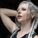 A woman with intricate braided white hair and silver hoop earrings poses with one arm raised behind her head. Sporting the Kaya Movement AYA Septum Clicker Shipibo Pattern in sterling silver 925 (16 gauge) and a black, cut-out top, she commands attention. The dark background accentuates her striking features prominently.