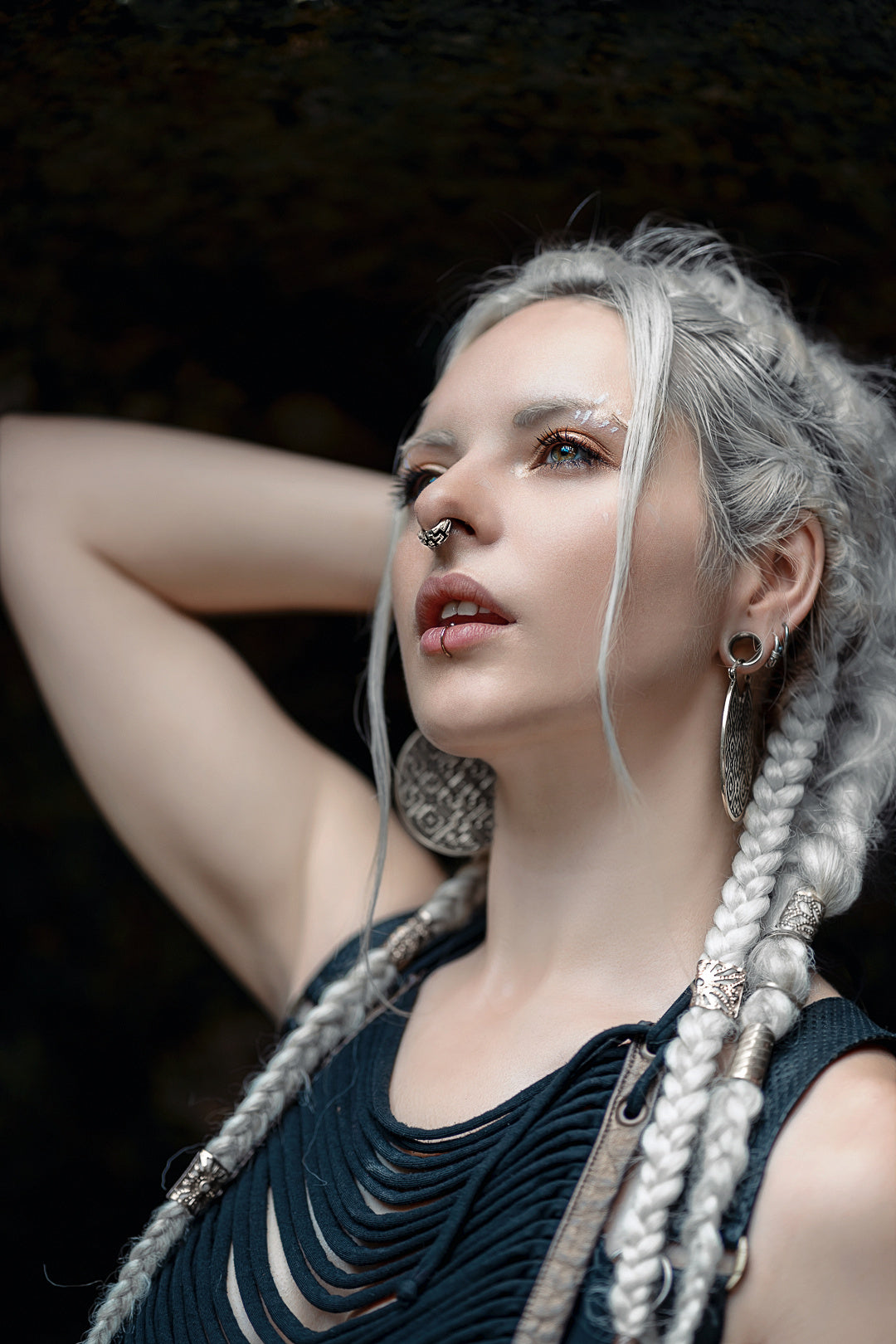 A woman with intricate braided white hair and silver hoop earrings poses with one arm raised behind her head. Sporting the Kaya Movement AYA Septum Clicker Shipibo Pattern in sterling silver 925 (16 gauge) and a black, cut-out top, she commands attention. The dark background accentuates her striking features prominently.
