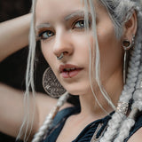Close-up of a person with long white braided hair, subtle makeup, and striking light-colored eyes. They have an AYA Septum Clicker Shipibo Pattern Inspired by Silver 925 from Kaya Movement (16 gauge), multiple earrings, and are wearing a dark, textured top. Their expression is serious as they gaze directly into the camera.