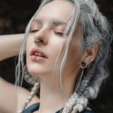 A person with long, braided white hair stands with eyes closed and one arm raised. They are wearing intricate sterling silver 925 earrings, an AYA Septum Clicker Shipibo Pattern Inspired by Silver 925 from Kaya Movement (16 gauge), and a lip piercing. The expression on their face appears serene. A dark and blurred background complements their appearance.
