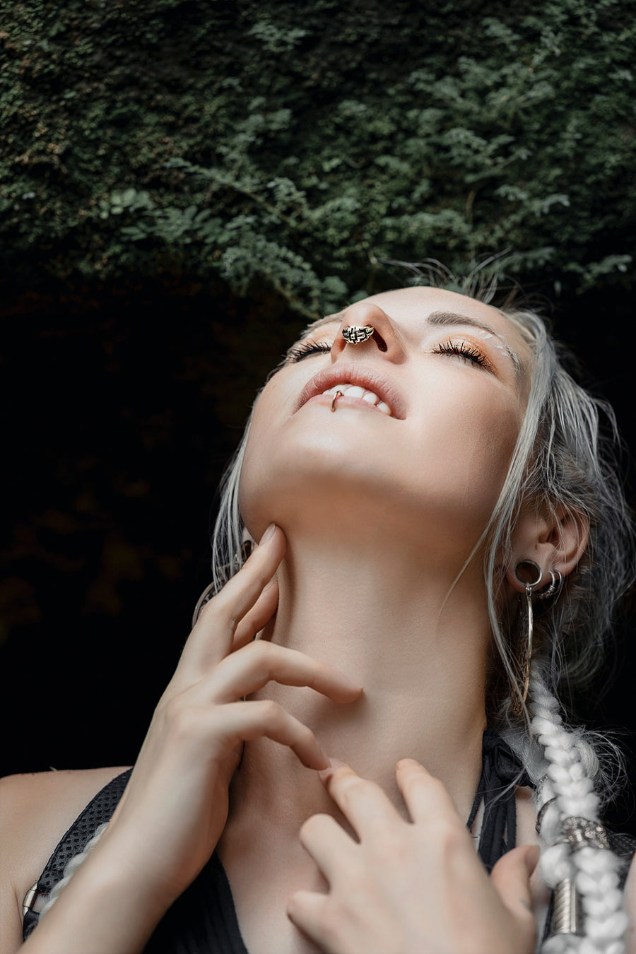 Contemplative woman gazing upward with detailed septum piercing and hoop earring, her hands gently touching her throat in a forest setting, highlighting a serene moment of connection with nature and self-expression through body jewelry.