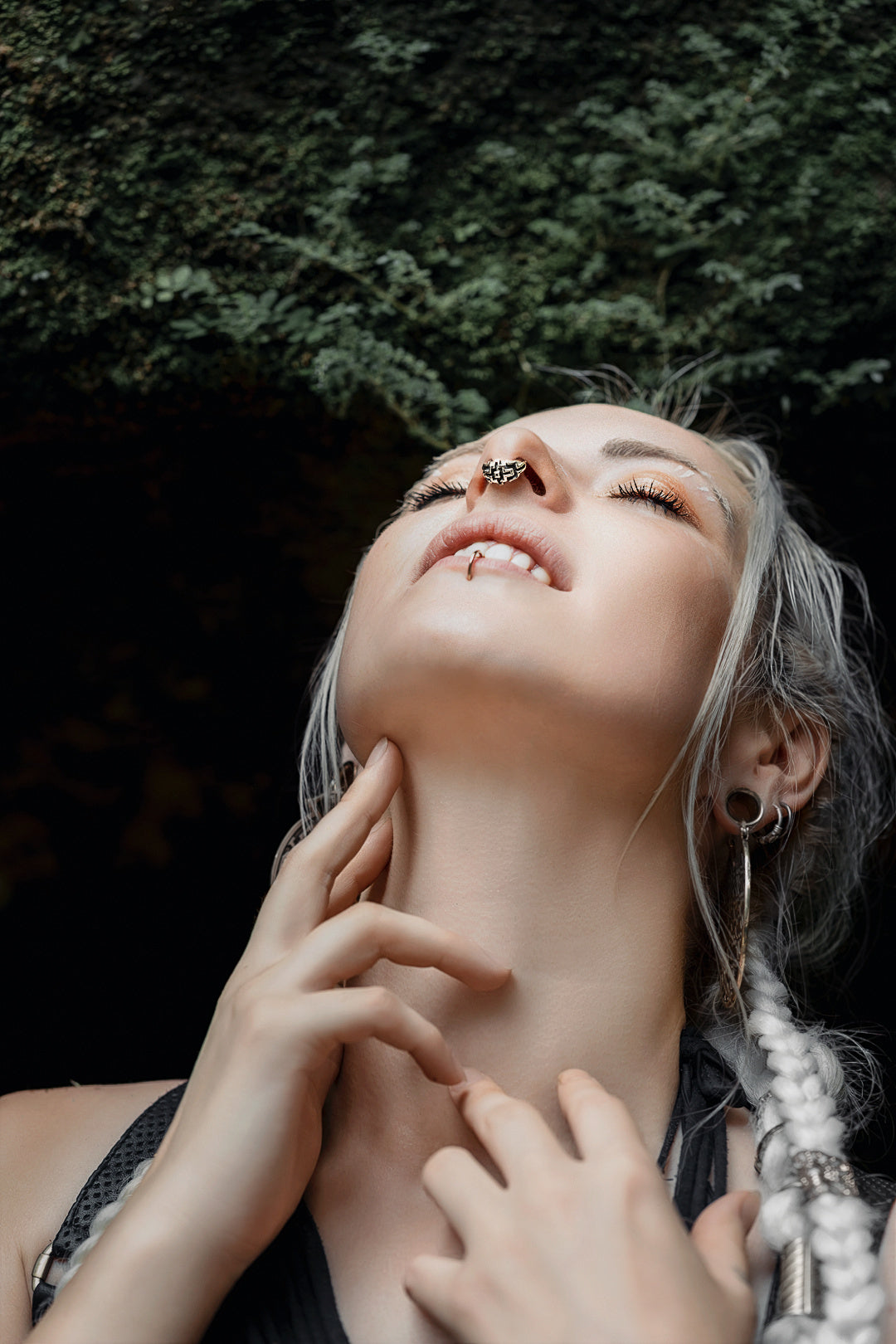 A person with braided hair and facial piercings, including the Kaya Movement AYA Septum Clicker Shipibo Pattern Inspired by Silver 925, tilts their head back, eyes closed, and smiles softly. They hold their neck with one hand and are set against a dark, natural background with greenery above.