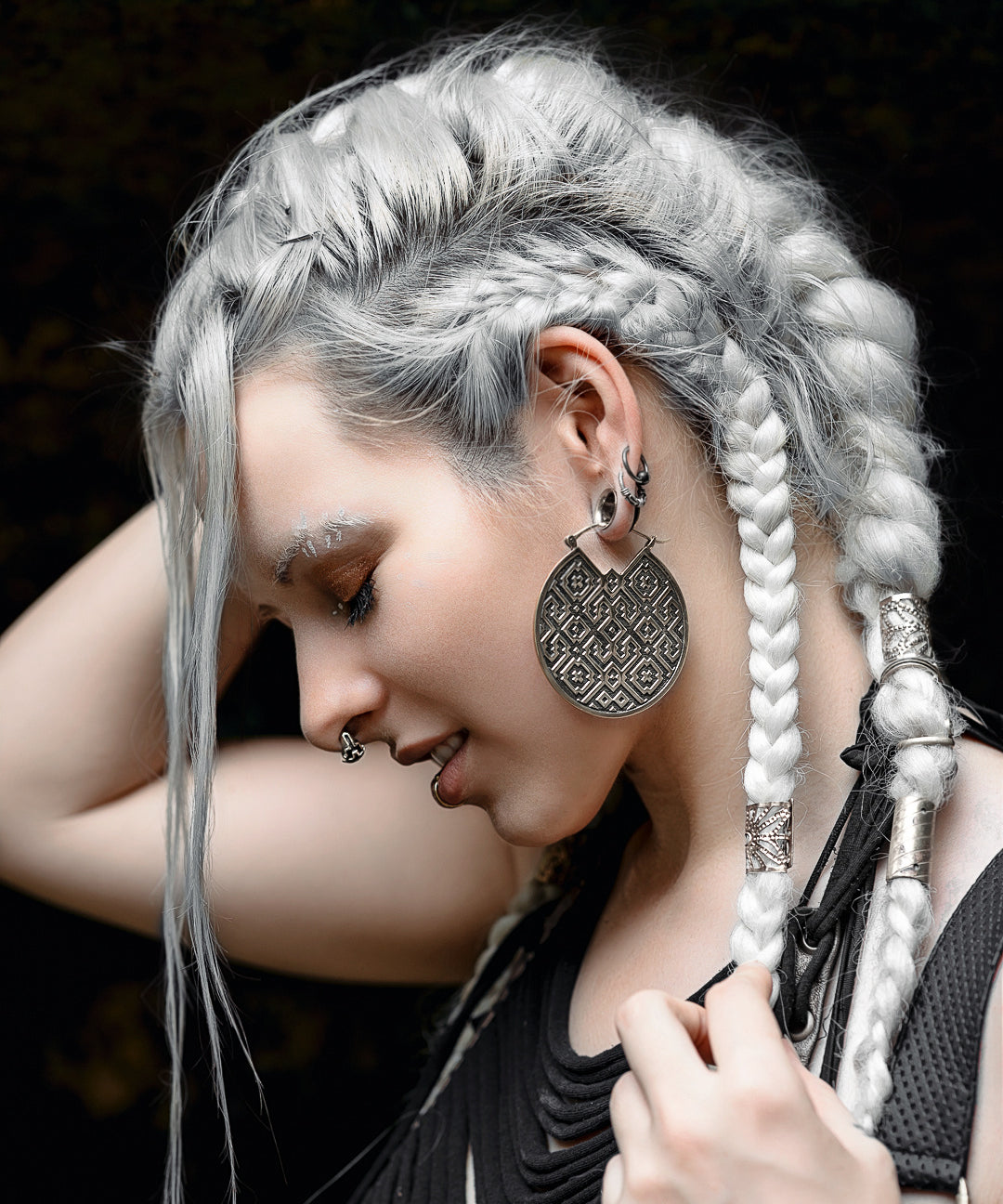 A person with long, platinum braided hair adorned with silver bands is shown in profile. They wear large, intricate circular earrings made from Sterling Silver 925 and have multiple facial piercings, including an AYA Septum Clicker Shipibo Pattern by Kaya Movement. Their hand gently touches their hair as they are dressed in black clothing.