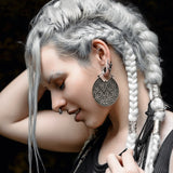 A person with long, platinum braided hair adorned with silver bands is shown in profile. They wear large, intricate circular earrings made from Sterling Silver 925 and have multiple facial piercings, including an AYA Septum Clicker Shipibo Pattern by Kaya Movement. Their hand gently touches their hair as they are dressed in black clothing.