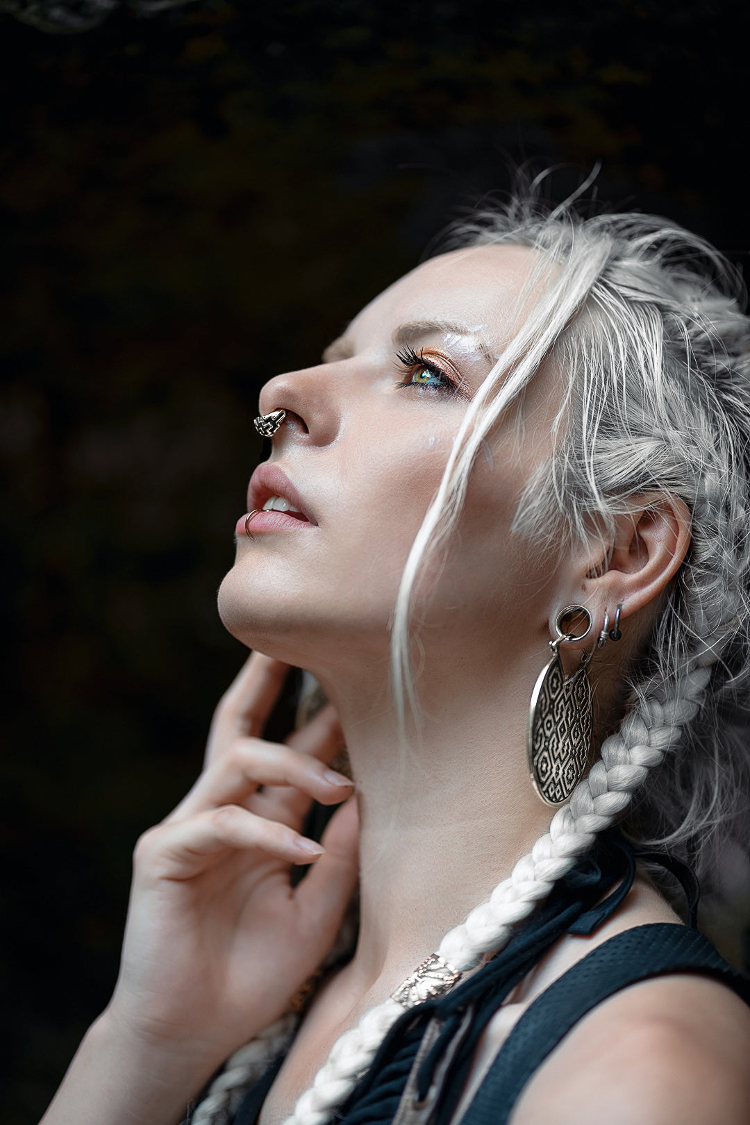 A person with pale skin and long, braided white hair looks upward. They have a Kaya Movement AYA Septum Clicker Shipibo Pattern Inspired by Silver 925 in a 16 gauge, a large earring, and are touching their chin with one hand. The background is dark and blurred, emphasizing their face and expression.