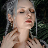 A person with long, braided gray hair is posed against a backdrop of green leaves. They have multiple piercings, including Kaya Movement’s KAMO Biomech Teardrop Septum Ring in Gold (16 gauge), a nose ring, and a lip ring. Their makeup includes shimmering eyeshadow. They are gently touching their neck with both hands.