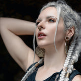 A woman with long white braids and silver jewelry poses with her arm behind her head, wearing a dark, intricately designed top featuring Shipibo patterns. She accessorizes with large circular earrings and the AYA Septum Clicker by Kaya Movement, inspired by icaros patterns in gold (16 gauge). Her eyes are accentuated with makeup against a dark and indistinct background.