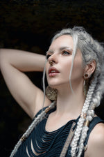 A woman with long white braids and silver jewelry poses with her arm behind her head, wearing a dark, intricately designed top featuring Shipibo patterns. She accessorizes with large circular earrings and the AYA Septum Clicker by Kaya Movement, inspired by icaros patterns in gold (16 gauge). Her eyes are accentuated with makeup against a dark and indistinct background.