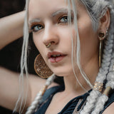 A person with long, white braided hair and piercing eyes gazes intently at the camera. They are wearing large, ornate earrings, a AYA Septum Clicker by Kaya Movement (Gold, 16 gauge) adorned with Shipibo pattern inspired by icaros, and a lip ring. The person holds one hand behind their head, revealing intricate braiding details and an embellished outfit showcasing Shipibo design influences.