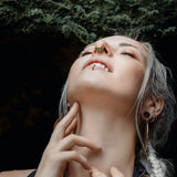 A woman with silver hair styled in braids looks upwards with her eyes closed. She adorns a Kaya Movement AYA Septum Clicker, inspired by Shipibo icaros patterns, a lip ring, and hoop earrings. Her fingers gently touch her neck and chest amidst a background of green, leafy foliage.