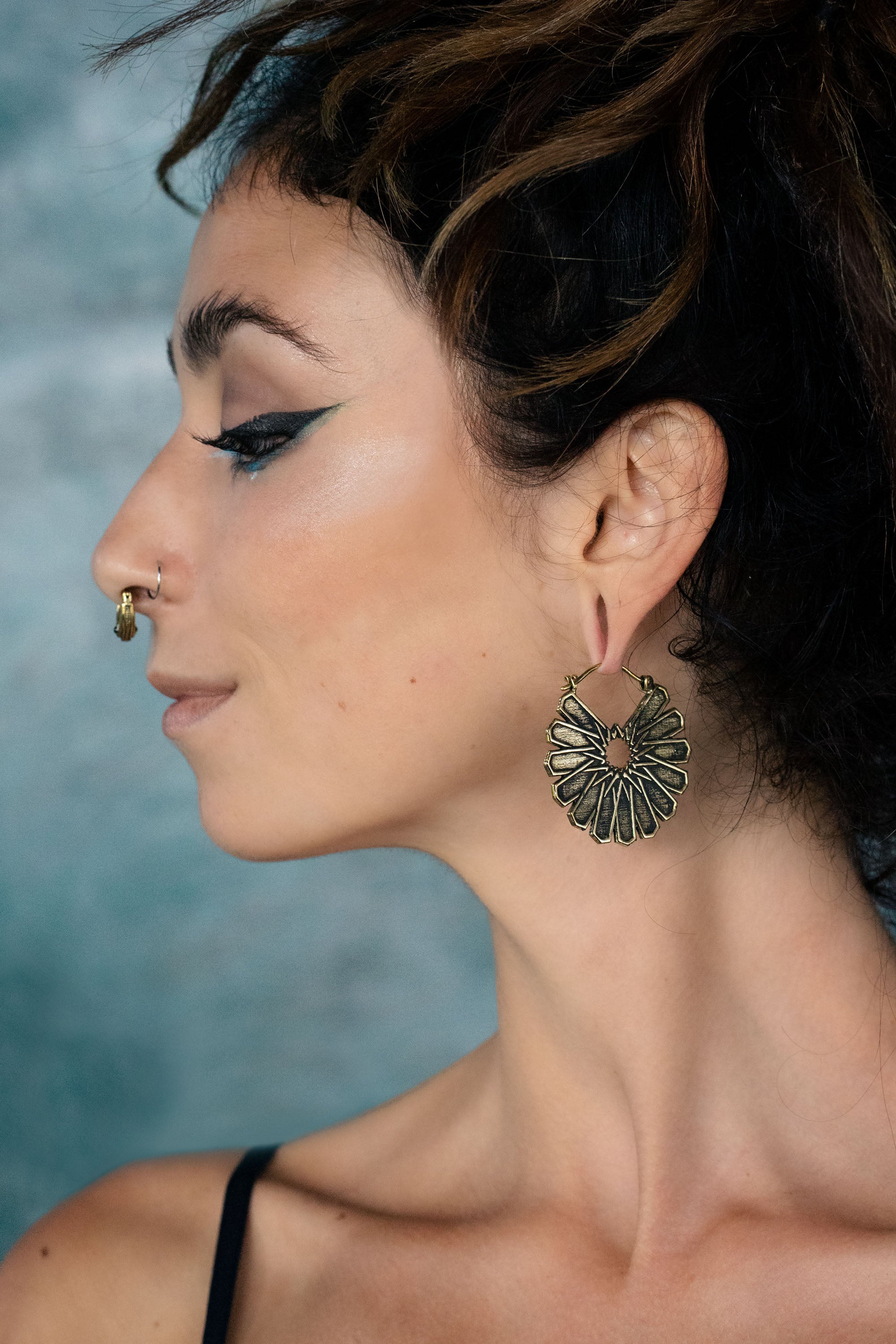 Side profile woman with an elaborate hairstyle and turquoise eyeliner, wearing gold septum clicker and a gold mozaic arabic earrings