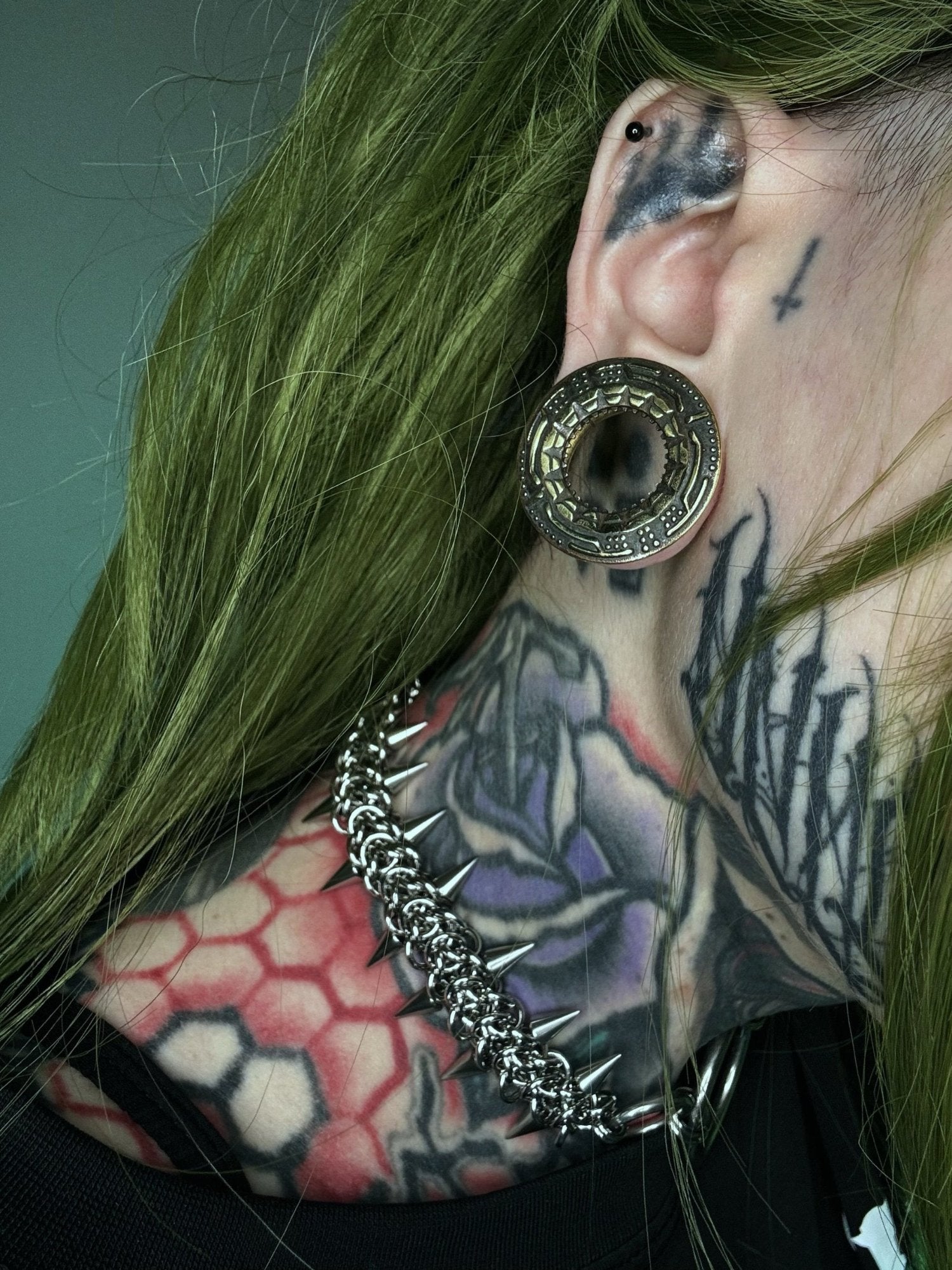 Close-up of a green-haired person with a large gold tunnel in a stretched ear, featuring an alien spaceship design. They have neck tattoos with geometric and floral patterns, and wear a spiked chain necklace, emphasizing a futuristic, cyberpunk style