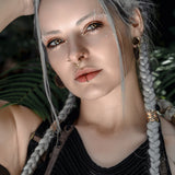 A person with long, braided silver hair and multiple facial piercings, including the KAMO Biomech Teardrop Septum Ring by Kaya Movement in gold, gazes intently at the camera. They are wearing a black, textured top and have their arm raised, touching their head. The background is blurred with green foliage.