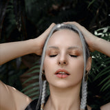 A woman with platinum blonde braids and minimalistic makeup holds her hands on the sides of her head. She wears the KAMO Biomech Teardrop Septum Ring in Gold by Kaya Movement (16 gauge) and a lip ring, both adorned with gold brass jewelry. Clad in a black top, she stands against lush green foliage, eyes closed and appearing serene.