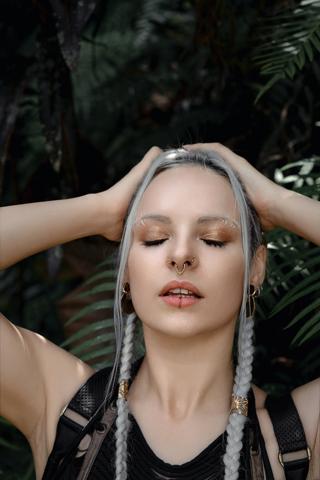 A woman with platinum blonde braids and minimalistic makeup holds her hands on the sides of her head. She wears the KAMO Biomech Teardrop Septum Ring in Gold by Kaya Movement (16 gauge) and a lip ring, both adorned with gold brass jewelry. Clad in a black top, she stands against lush green foliage, eyes closed and appearing serene.