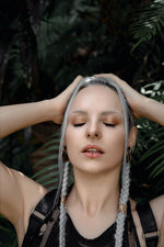 A woman with platinum blonde braids and minimalistic makeup holds her hands on the sides of her head. She wears the KAMO Biomech Teardrop Septum Ring in Gold by Kaya Movement (16 gauge) and a lip ring, both adorned with gold brass jewelry. Clad in a black top, she stands against lush green foliage, eyes closed and appearing serene.