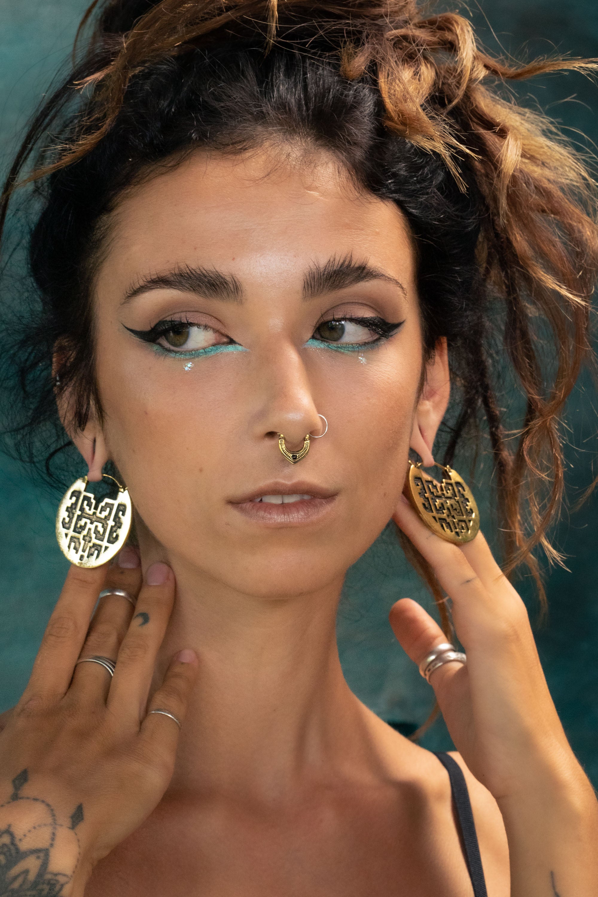 Woman with an elaborate hairstyle and turquoise eyeliner, wearing a gold septum ring and large round golden earrings with geometric cut-out designs. Her hands frame her face, showcasing rings and a mandala-style tattoo on her wrist.