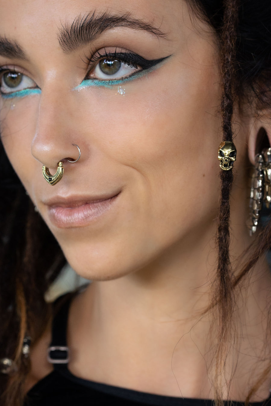 Artistic side portrait of a woman with bold winged eyeliner and turquoise under-eye accents, wearing a gold septum ring and a gold skull bead in her dreadlock. She smiles softly, showing a confident and playful vibe.